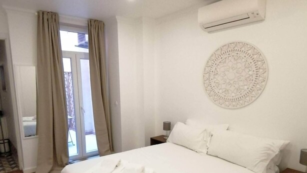 Quarto de hotel com cama, roupa de cama branca, unidade de ar condicionado, um objeto decorativo na parede e vista por uma porta.