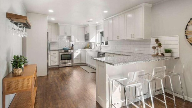 A modern kitchen with white cabinets, a central island with bar stools, and a wooden dining table with a plant. - BA's BohoChic-steps to Rose District w/Hot Tub (Broken Arrow)