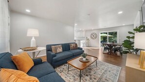 A modern living room with a blue sofa, a wooden coffee table, and a dining area with a large window. - BA's BohoChic-steps to Rose District w/Hot Tub (Broken Arrow)
