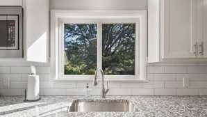 A kitchen with a granite countertop, stainless steel sink, and a window with a view of trees. - BA's BohoChic-steps to Rose District w/Hot Tub (Broken Arrow)