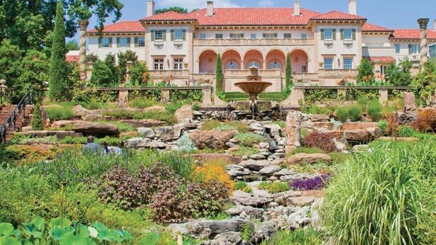 A large, multi-story building with a red-tiled roof, a central courtyard with a fountain, and a well-maintained garden with various plants and rocks. - Ranch Acres Expo-Heart of Tulsa (Tulsa)