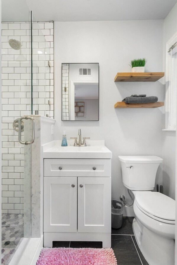 Une salle de bain avec une douche à parois de verre, des carreaux blancs, un lavabo blanc avec un miroir et des toilettes.