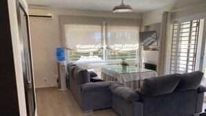 Un salon moderne avec un canapé gris, une table à plateau en verre et une vue sur une piscine, visible à travers une fenêtre ornée de rideaux légers.