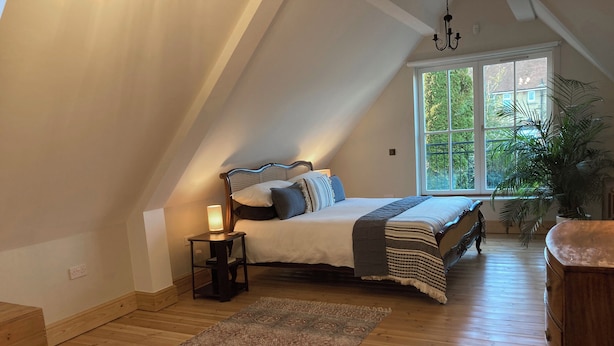 A bedroom with a sloped ceiling, a large window, a bed with a headboard, a bedside table with a lamp, and a wooden floor. - Gingerbread Cottage (Canterbury)