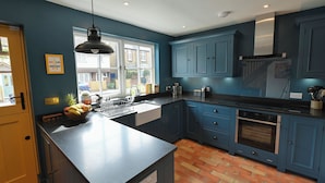 A kitchen with blue cabinets, a black countertop, a window with a view of the outside, and a brick-patterned floor. - Gingerbread Cottage (Canterbury)