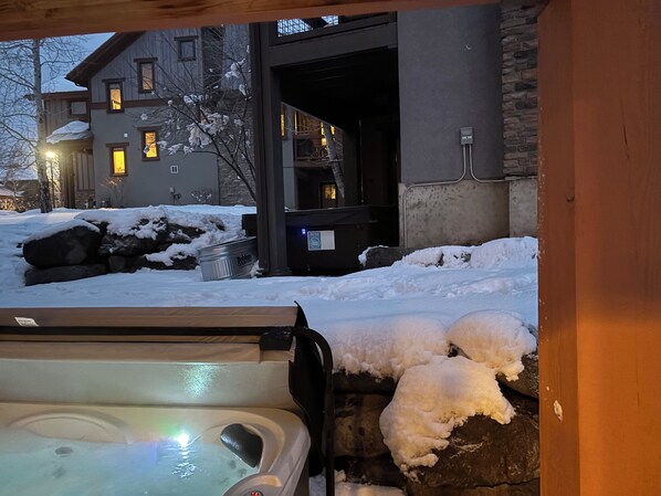 A hot tub with snow on its exterior, a stone wall, and a building with windows in the background.