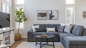 A modern living room with a grey sectional sofa, a small round coffee table, a TV on a white cabinet, and a large potted plant. - Perfect Family Haven: 5BR,Heated Pool + Golf Green (Gilbert)