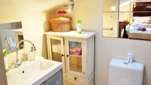 Casa de banho com lavatório, espelho e sanita. Há uma cesta de vime sobre o balcão do lavatório e um rolo de papel higiénico no armário.