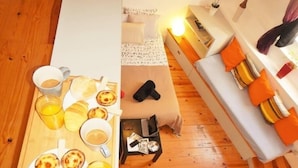 Um quarto acolhedor com piso de madeira, uma cama com almofadas laranja e brancas, um sofá com almofadas laranja e uma bandeja de café da manhã com pães e bebidas.