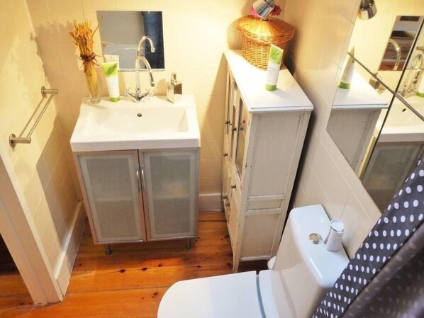 Une petite salle de bain avec un lavabo blanc, un miroir et des toilettes. On y trouve un panier en osier sur le comptoir et une bouteille de lotion.