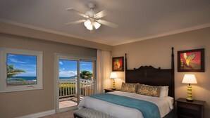Une chambre à coucher avec un lit, deux tables de chevet avec des lampes, un banc, un ventilateur de plafond, une vue sur l’océan par une fenêtre et des œuvres d’art accrochées aux murs.