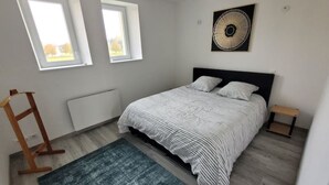 A bedroom with a bed, a standing heater, a small table, and two windows.