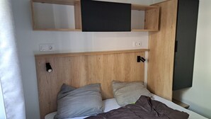 A bedroom with a wooden headboard, a bed with grey and brown bedding, and a flat-screen TV mounted on the wall. - NEW AIR CONDITIONED MOBIL HOME - CAMPING 5* - FOR 6 PEOPLE with dishwasher (Lagorce)