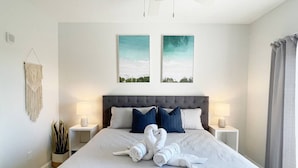 Una cama bien hecha con un cabecero gris, dos lámparas de noche y un tapiz de macramé en la pared.