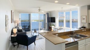 Un apartamento moderno con sala de estar, comedor y cocina, con ventanas amplias y un balcón con vista a un cuerpo de agua.