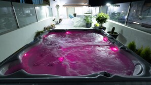 Uma banheira de hidromassagem iluminada em rosa, rodeada por plantas em vasos e uma pequena área de estar externa.