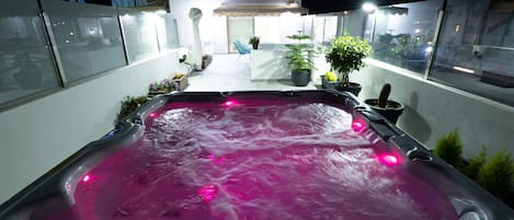 Ein Whirlpool mit rosarotem Licht, umgeben von Blumentöpfen und einer kleinen Sitzgruppe im Freien.