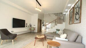 Uma sala de estar moderna com um sofá cinzento, uma poltrona escura, uma mesa de centro de madeira e uma TV fixada na parede.