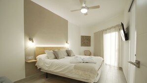 Quarto com cama, mesas de cabeceira, ventilador de teto, televisão fixada na parede e uma janela grande com cortinas.