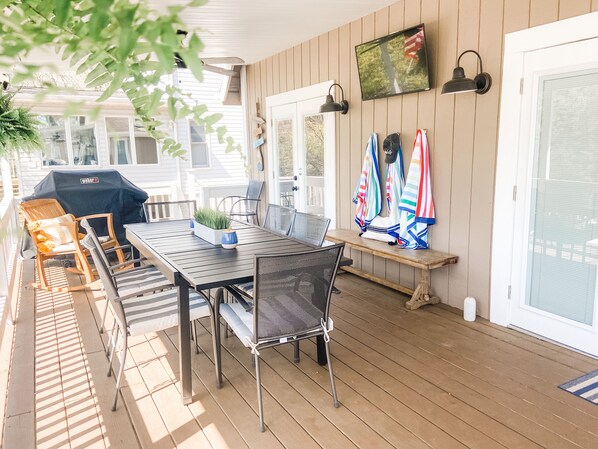 Eine überdachte Terrasse mit Tisch, Stühlen, einem Grill und einem an der Wand befestigten Fernseher.