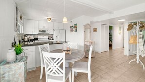 A modern kitchen with white cabinetry, a central island, and pendant lighting. There is a dining area with a round table and white chairs. - Cocoa Beach Direct Oceanfront Condo - just a few steps to the beach! (Cocoa Beach)