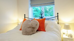 A neatly made bed with white and orange bedding, a window with a view of greenery, and a bedside table with a lamp and a decorative item.