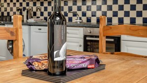 A bottle of wine, a chocolate bar, and a stack of books on a wooden table in a kitchen with checkered tiles and white cabinets. - Robin’s Nest - UK36208 (Capel Seion, near Aberystwyth)
