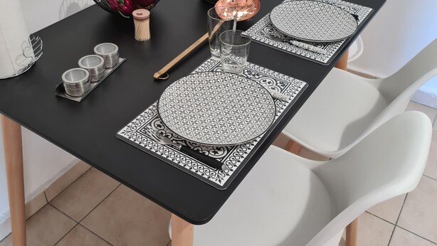 Una mesa de comedor con manteles individuales blancos y negros, una olla de cobre, un frutero y sillas blancas.