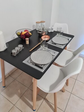 Une table à manger dressée avec des sets de table à motifs noirs et blancs, un pot en cuivre, un saladier rempli de fruits et des chaises blanches.