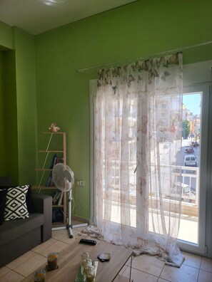 A living room with a green wall, a sofa, a fan, a table with drinks, and a window with a curtain.