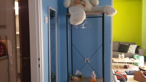 Una habitación con pared azul, un oso de peluche blanco sobre una estantería de metal negro, una pequeña mesa de madera con objetos encima y una pared verde al fondo.