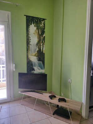 Une pièce avec un mur vert, une grande affiche représentant une cascade, un téléviseur à écran plat posé sur un meuble en bois, et deux manettes de jeu sur le meuble.