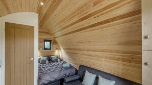 Un dormitorio con techo inclinado, paredes de madera, cama con cubrecama oscuro, sofá gris con cojines y ventana con vista a árboles.
