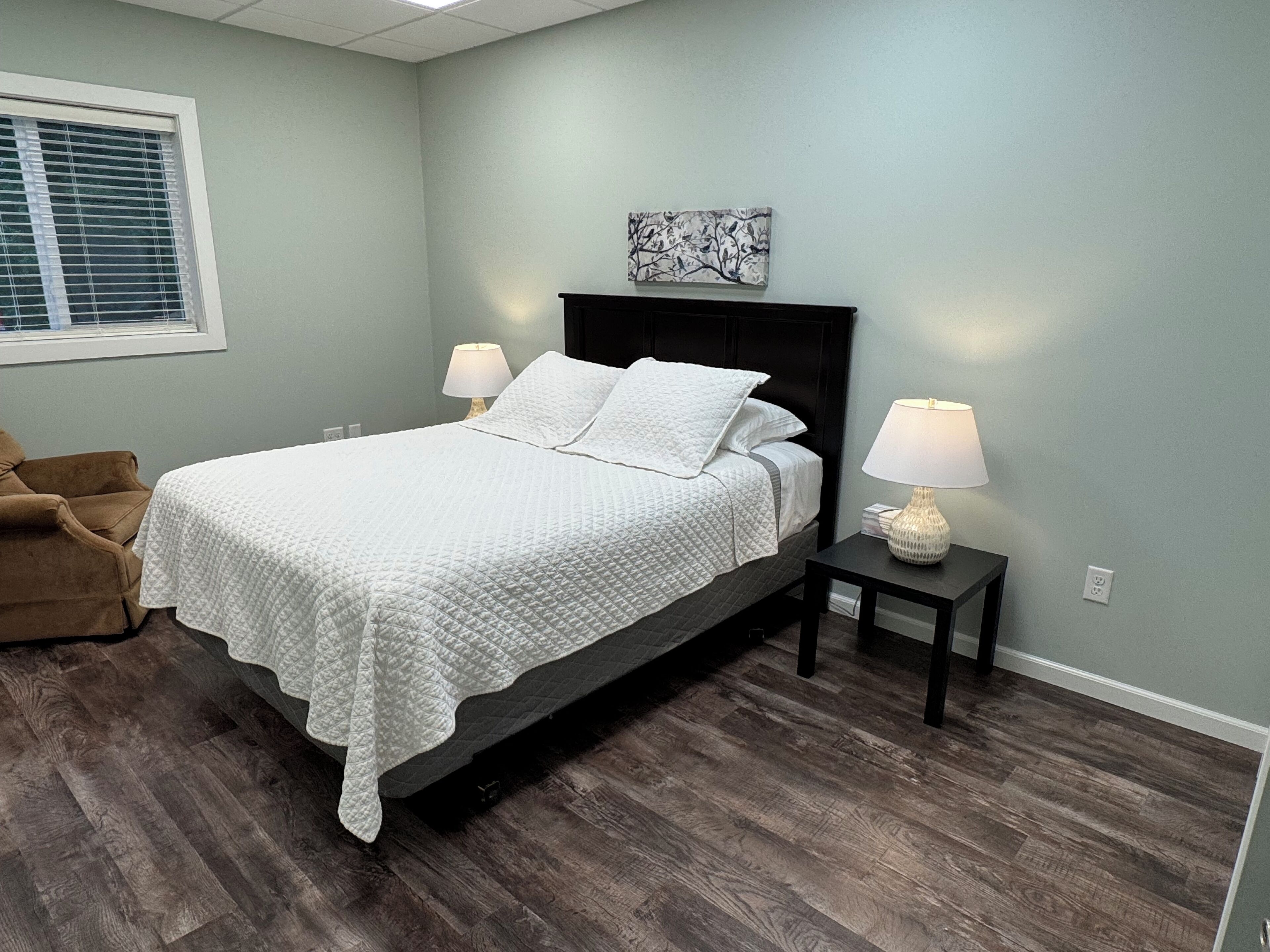 Une chambre à coucher comprenant un lit, deux tables de chevet, une commode, une chaise et une fenêtre avec des stores.
