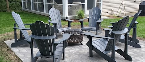 Un patio con sillas Adirondack y una fogata, un porche cubierto y una casa al fondo.