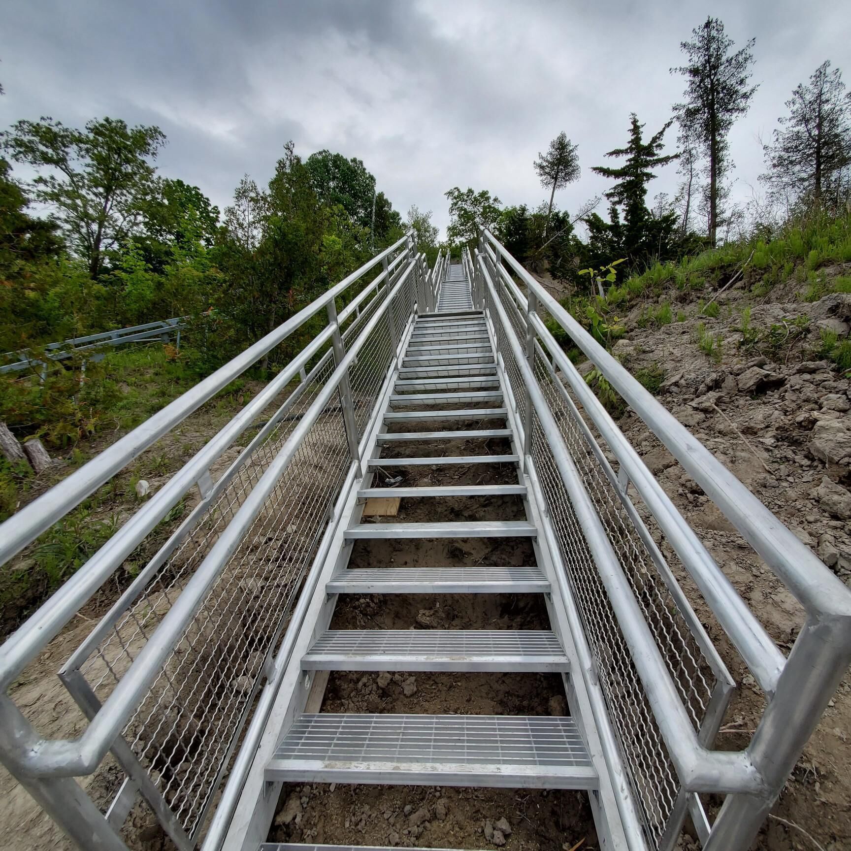Un escalier métallique avec des rampes menant vers le haut, entouré de verdure et d’arbres.