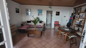 Een woonkamer met een bank, een salontafel en een eettafel met stoelen. Er is een deur naar een andere kamer en een plank met diverse spullen.