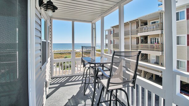 A balcony with a table and chairs, overlooking a beach and adjacent buildings. - Oceanfront Windsong Unit 207 | 1BR/1BA | Sleeps 5 | Queen, Bunks, Sofa Beds Setu (North Myrtle Beach)