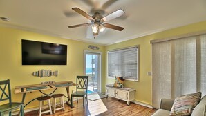A living room with a wooden floor, a ceiling fan, a flat-screen TV, a dining table with chairs, a sofa, and a window with blinds. - Oceanfront Windsong Unit 207 | 1BR/1BA | Sleeps 5 | Queen, Bunks, Sofa Beds Setu (North Myrtle Beach)