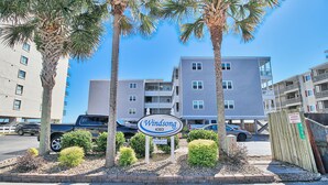 A sign for Windsong 4303, a multi-story building with balconies, surrounded by palm trees and a parking area. - Oceanfront Windsong Unit 207 | 1BR/1BA | Sleeps 5 | Queen, Bunks, Sofa Beds Setu (North Myrtle Beach)