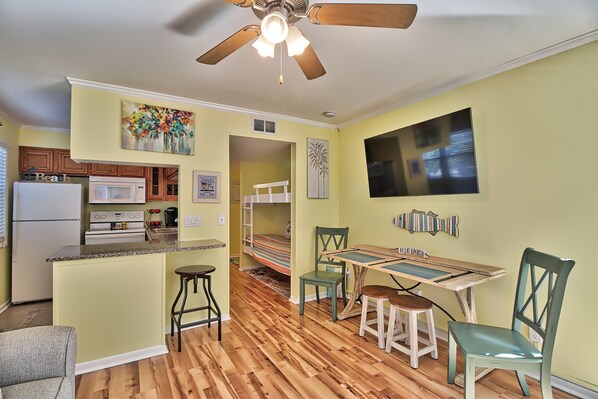 A compact kitchen with a refrigerator, microwave, and stove. There is a dining area with a table and chairs, and a bunk bed in the background. - Oceanfront Windsong Unit 207 | 1BR/1BA | Sleeps 5 | Queen, Bunks, Sofa Beds Setu (North Myrtle Beach)