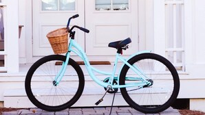 Una bicicleta turquesa estacionada frente a una puerta blanca con una cesta en el manillar.