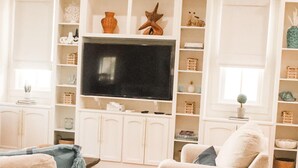 Una sala de estar con un televisor de pantalla plana grande, dos sofás, un taburete de madera pequeño y estanterías empotradas blancas con varios objetos decorativos.