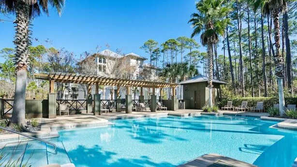 Una piscina con pérgola y sillas de descanso, rodeada de palmeras y un edificio al fondo.
