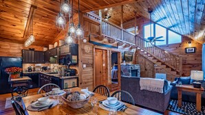 A wooden cabin interior with a dining area, kitchen, and a staircase leading to a loft.