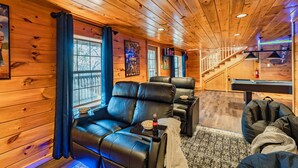 A cozy room with wooden walls and ceiling, a black leather sofa, a small table with a bottle, and a pool table.