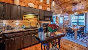 A wooden cabin interior with a kitchen, dining area, and a large window.