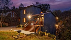 A two-story log cabin with a wooden staircase leading to a porch, surrounded by trees.