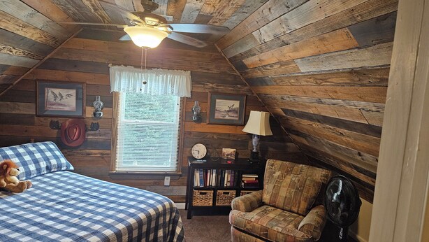 A cozy attic room with wooden walls, a ceiling fan, a large window with blinds, a bookshelf, and a comfortable seating area with a couch and a chair. - EXCLUSIVE LARGE 2 BEDROOM SUITE W/THEATER ROOM + KITCHEN  (Cleveland)