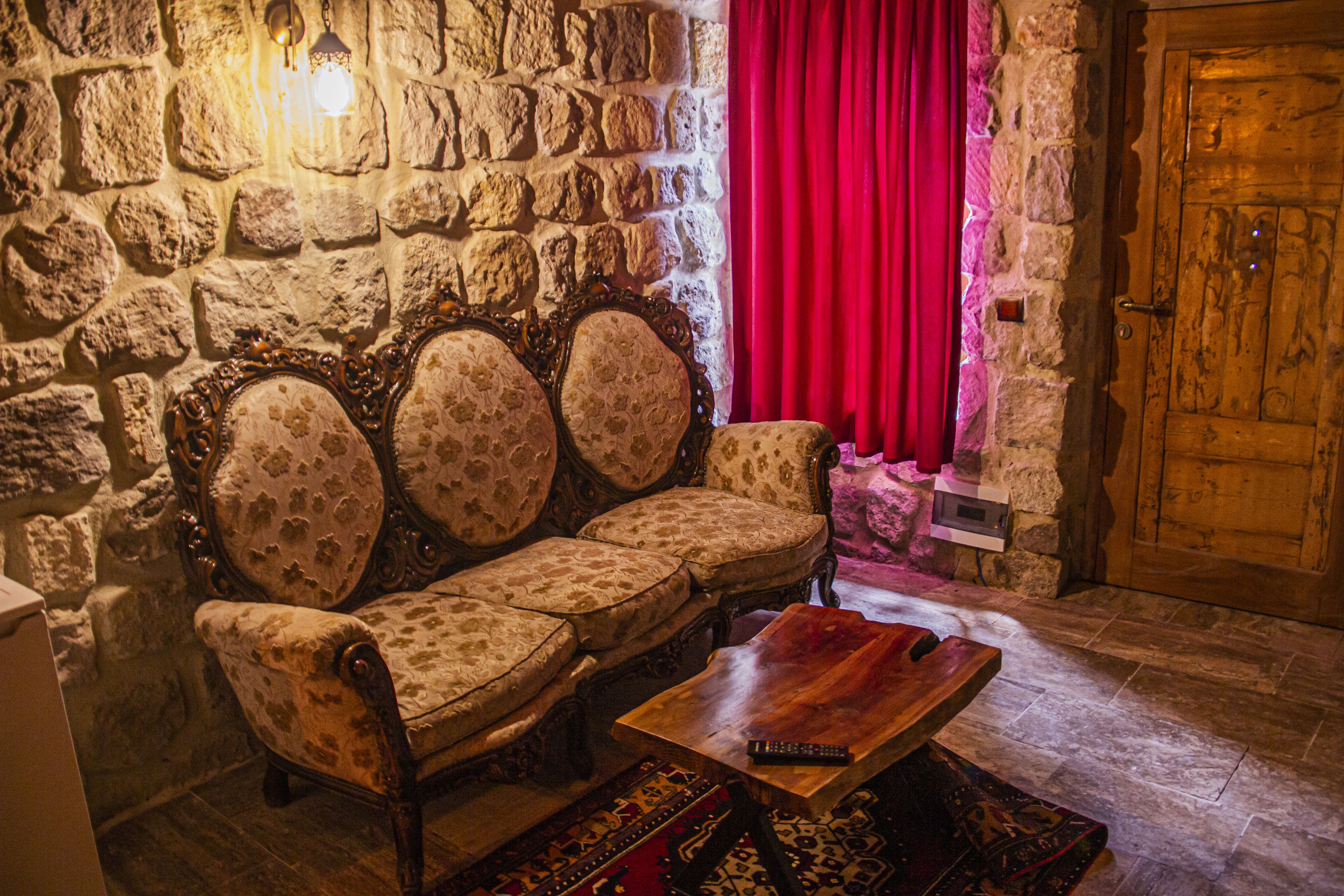 Una habitación con paredes de piedra, una puerta de madera, una cortina roja y un sofá antiguo con un control remoto sobre una mesa de centro de madera.